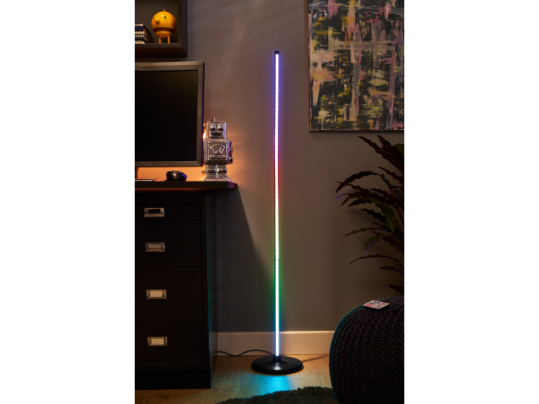 Une grande lampe de sol LED à couleurs changeantes se tient à côté d'un bureau sombre avec un moniteur d'ordinateur.