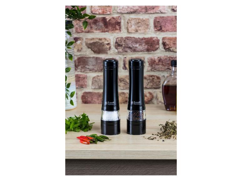 Deux moulins électriques noirs à sel et poivre sur un comptoir en bois, avec des herbes et des piments.
