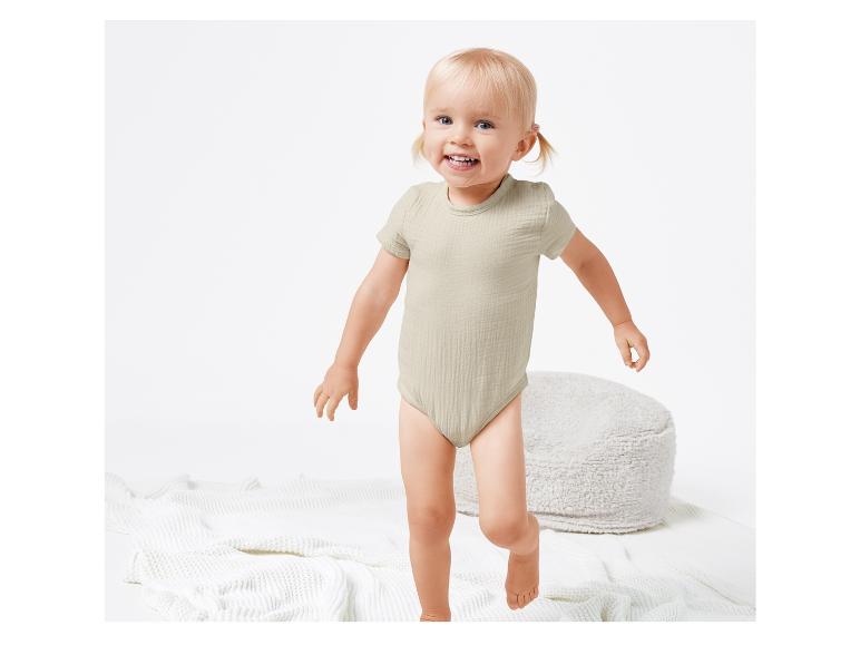 Bébé souriant portant un body beige et jouant sur un tapis blanc.