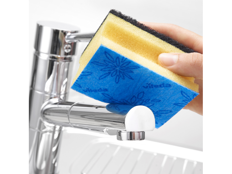 Main nettoyant un robinet de cuisine avec une éponge de nettoyage jaune et bleue.