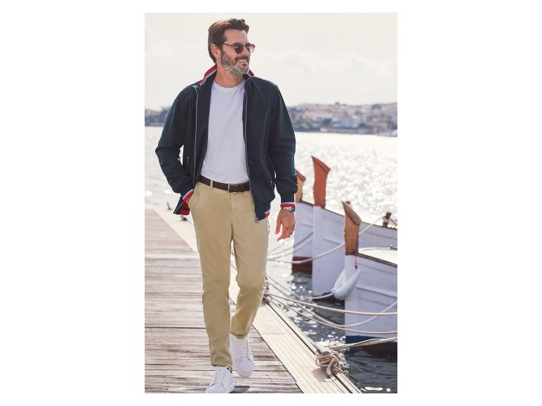 Un homme en veste bleue, pantalon beige et baskets blanches marche sur une jetée en bois.