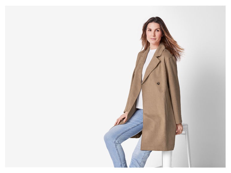 Femme en manteau brun et jeans posant près d'un tabouret blanc.