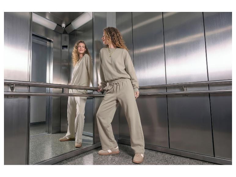 Femme en sweat-shirt et pantalon de survêtement beige, debout dans un ascenseur.