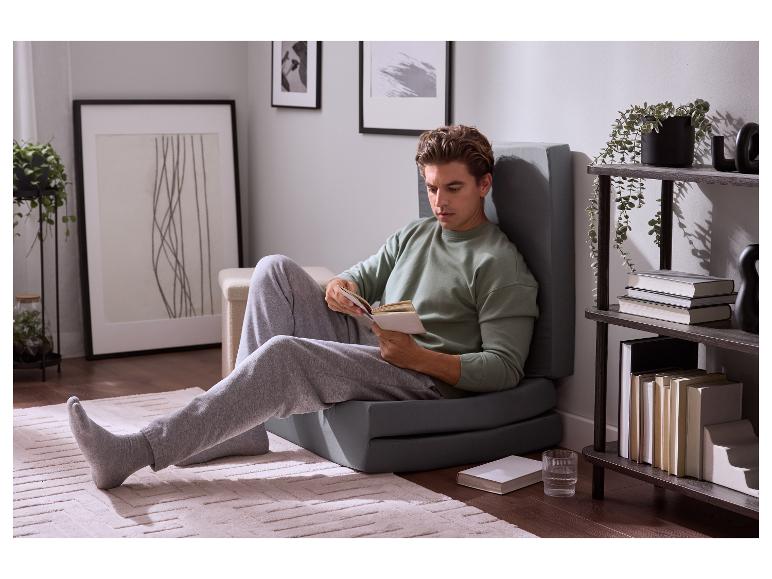 Homme lisant un livre, assis sur un matelas pliable gris dans un salon.