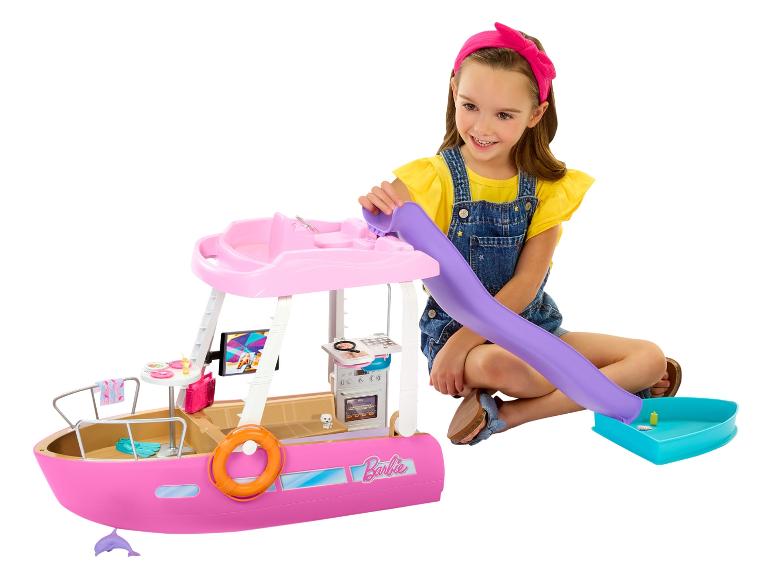 Une fille souriante joue avec un bateau jouet Barbie rose, avec toboggan et accessoires.