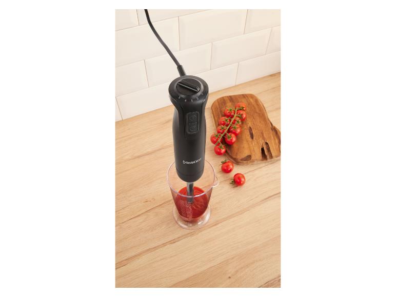 Mixeur plongeant dans un verre doseur avec sauce tomate, tomates cerises sur planche en bois