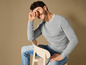 Un homme en pull gris et jean bleu, assis sur une chaise en bois et riant.