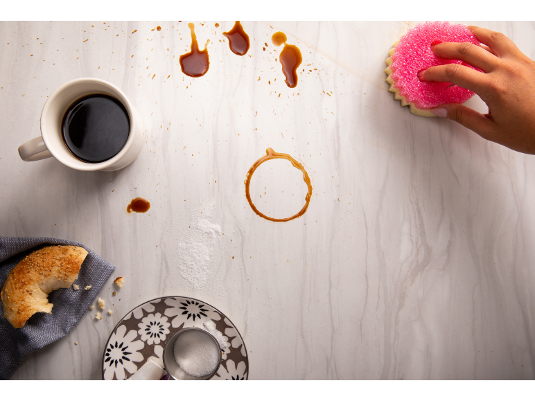 Une main nettoie une table avec une éponge, du café renversé, un bagel et du sucre.