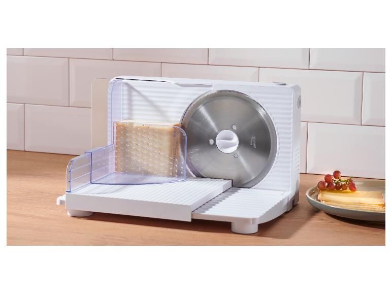 Une trancheuse à aliments blanche avec un bloc de fromage et une assiette de fromage tranché et de raisins.