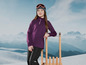 Une fille portant un pull violet et un pantalon noir s'appuie sur une luge en bois, devant un paysage montagneux.