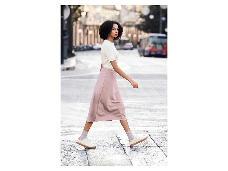 Une femme avec un haut blanc et une jupe rose marche dans la rue avec des baskets blanches.