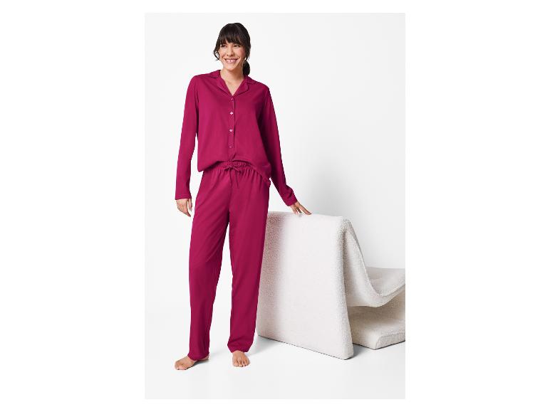 Femme en pyjama rouge foncé à manches longues et pantalon, debout près d'une chaise blanche.