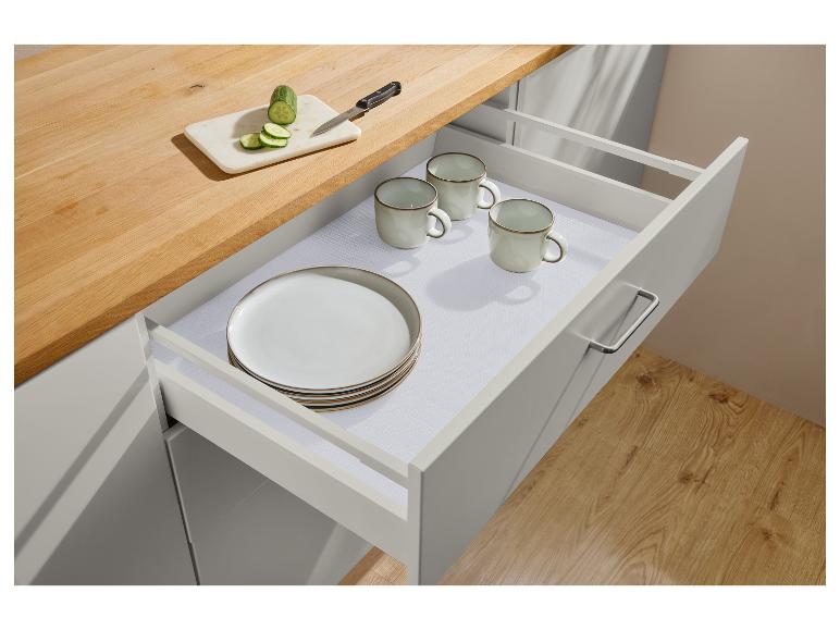 Tiroir de cuisine ouvert avec assiettes, tasses et tapis antidérapant, avec concombre coupé sur le comptoir.