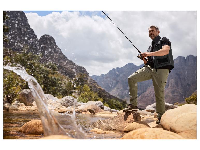 Homme pêchant dans une rivière, portant un gilet de pêche, un t-shirt et un pantalon cargo.
