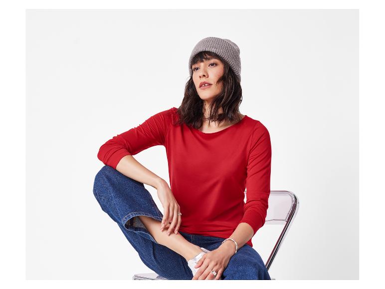 Femme en haut rouge à manches longues, jean bleu et bonnet gris assise sur une chaise.