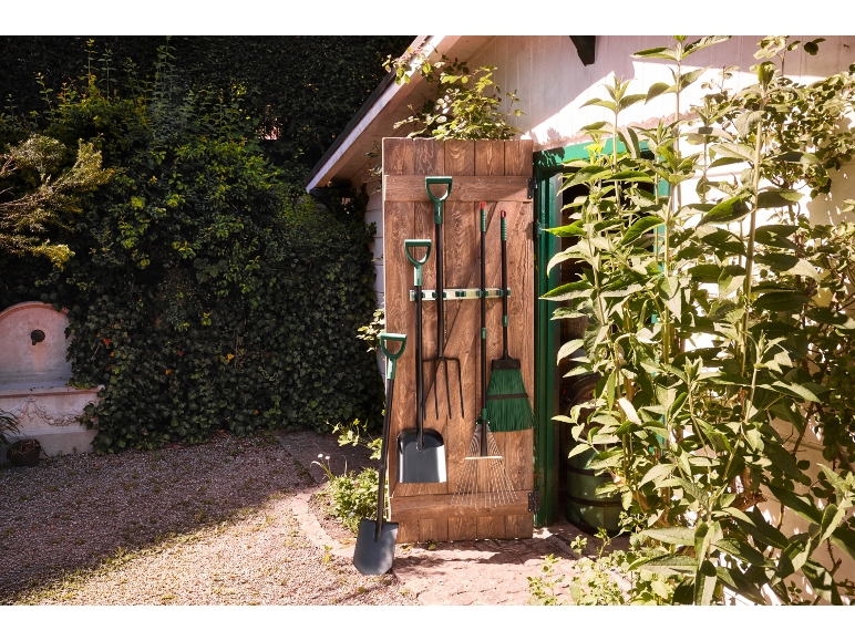 Outils de jardinage suspendus à une porte en bois, avec une pelle et un râteau au premier plan.