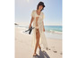 Une femme marche sur la plage en robe de chambre blanche, avec des tongs à la main.
