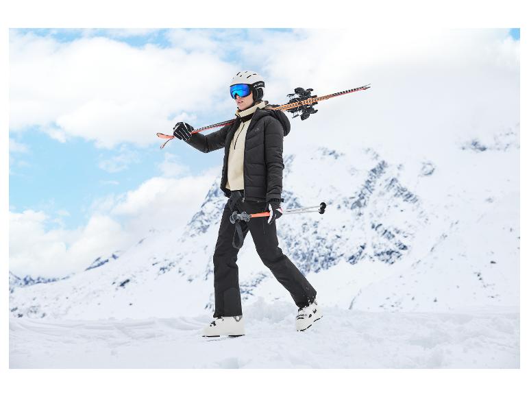 Femme en équipement de ski noir et blanc, skis sur l'épaule, sur une montagne enneigée.