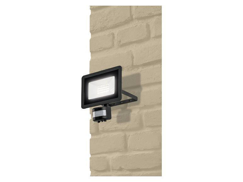 Projecteur LED noir avec détecteur de mouvement, monté sur un mur de briques beiges.