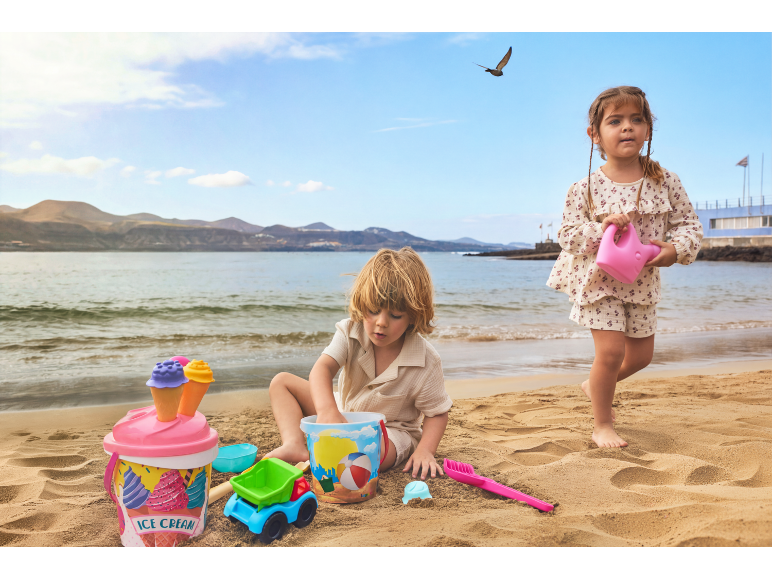 Deux enfants jouent avec des jouets de sable, dont un seau à glace et un arrosoir, sur la plage.