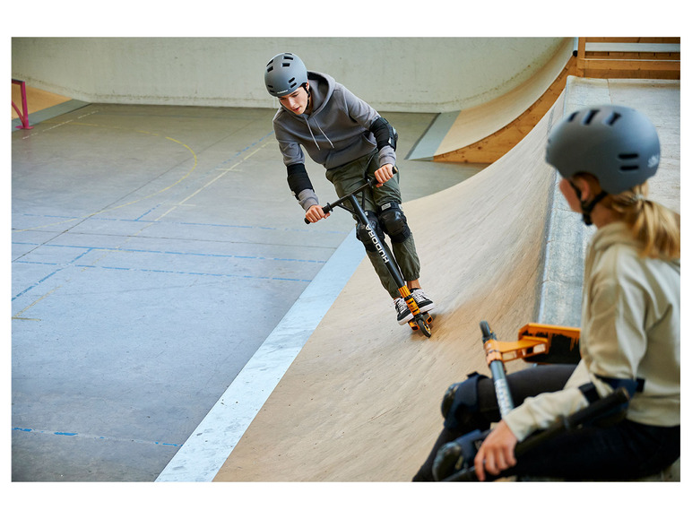 Deux personnes sur un skatepark, l'une sur une trottinette Hudora et l'autre sur un scooter.