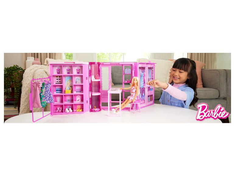 Fille jouant avec une garde-robe de poupée rose, une poupée et des accessoires sur une table blanche.