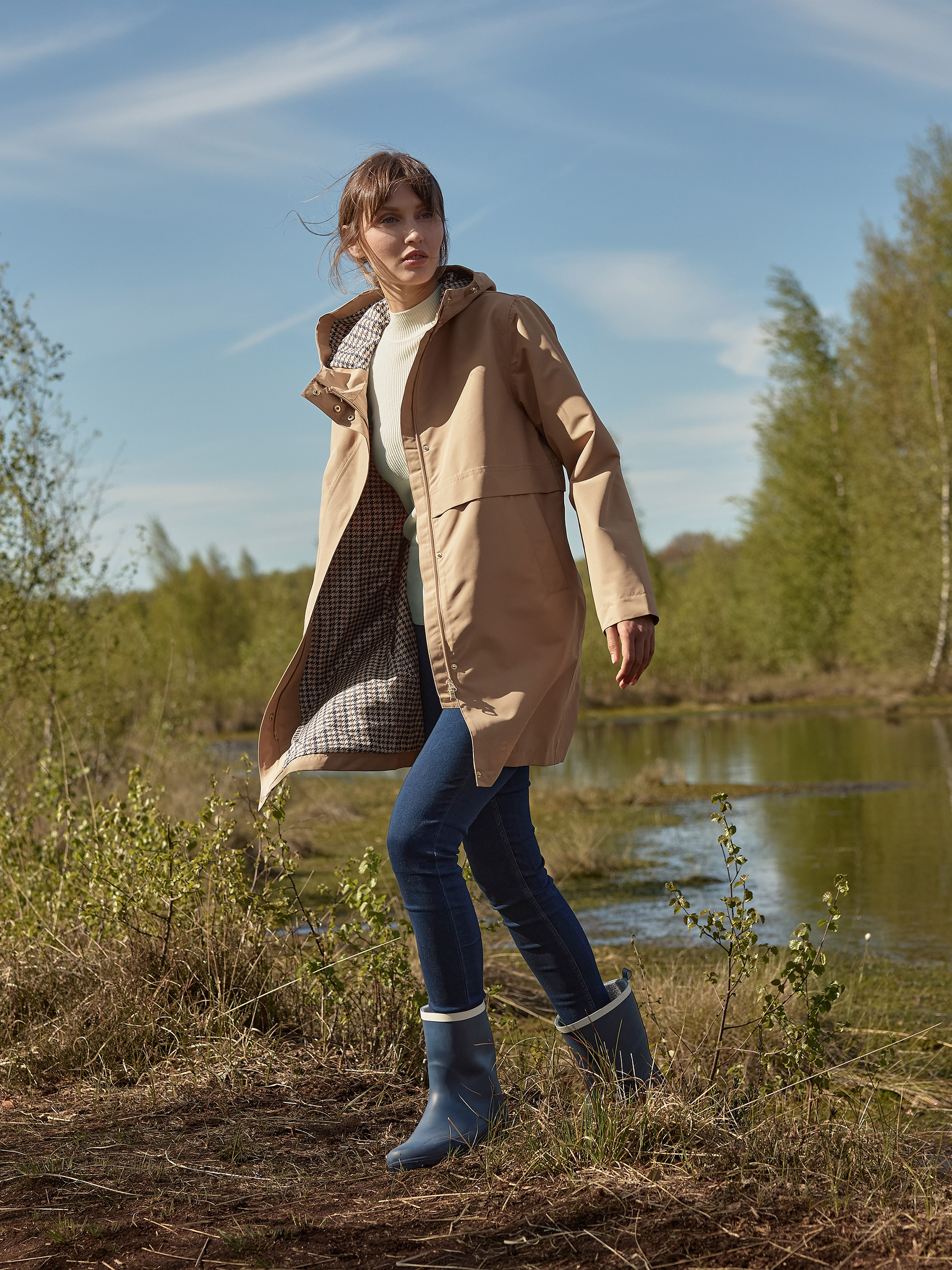 Femme en trench-coat beige, pull à col roulé, jean et bottes de pluie bleues, marchant près d'un lac.