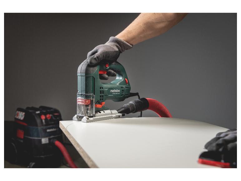 Scie sauteuse Metabo coupant du bois, connectée à un aspirateur, main gantée.