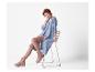 Femme rousse en long cardigan bleu et pantalon marron, assise sur une chaise.