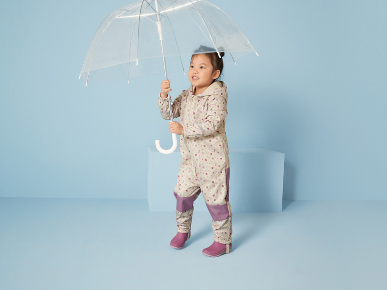 Un enfant avec une combinaison de pluie et des bottes tient un parapluie transparent.