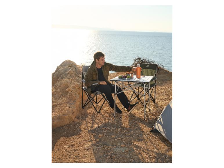 Homme en veste kaki assis sur une chaise de camping Crivit, près d'une table de camping avec une bouteille d'eau orange, face à la mer.