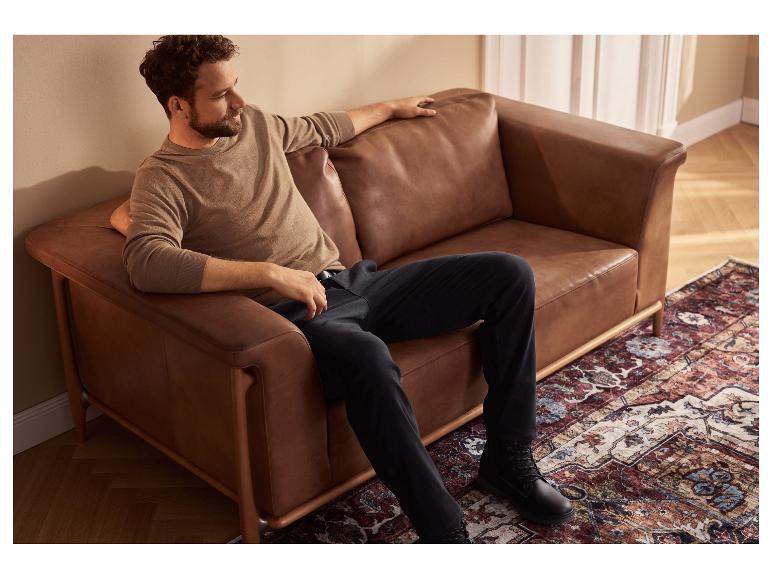 Un homme est assis sur un canapé en cuir marron, portant un pull beige et un pantalon noir.