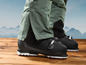 Des chaussures de ski noires avec un pantalon vert sur un plancher en bois.