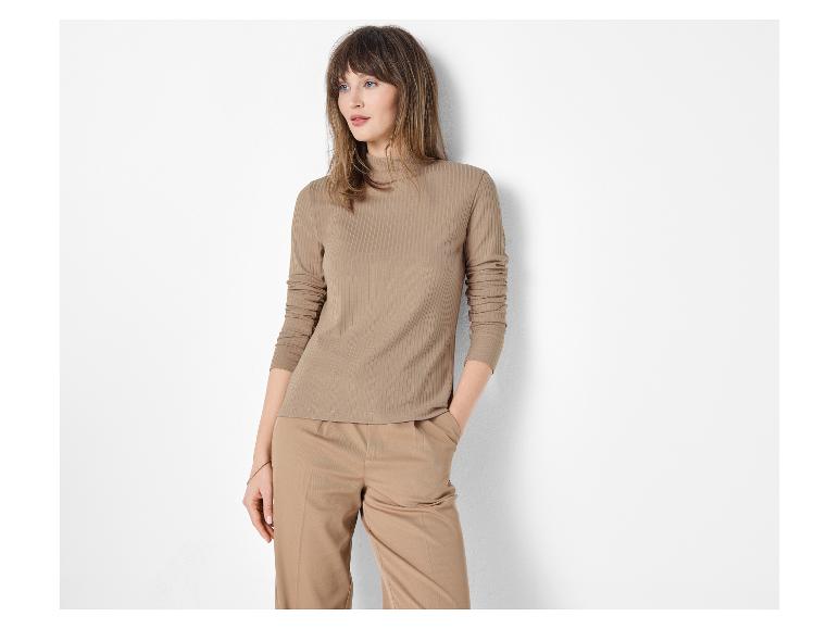 Femme en haut côtelé marron et pantalon marron posant contre un mur blanc.