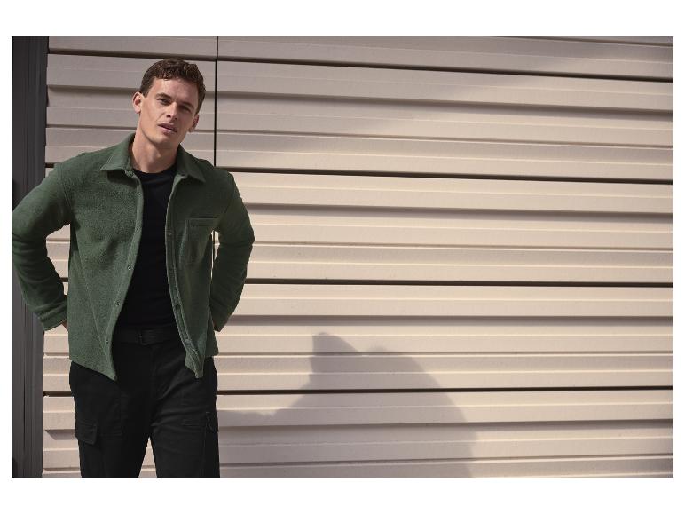 Homme en chemise polaire verte et pantalon cargo noir, posant devant un mur à lattes.