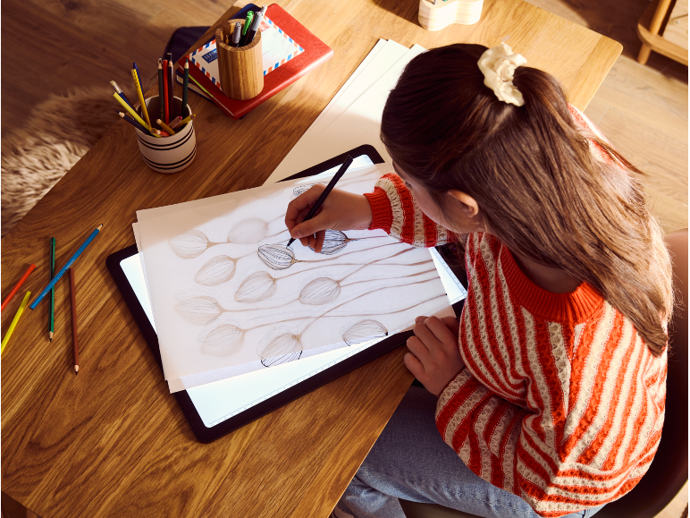 Femme dessinant sur une table lumineuse avec des crayons de couleur et des carnets sur une table en bois.