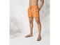 Un homme porte un short de bain orange avec un motif de crabes et des tongs bleues.