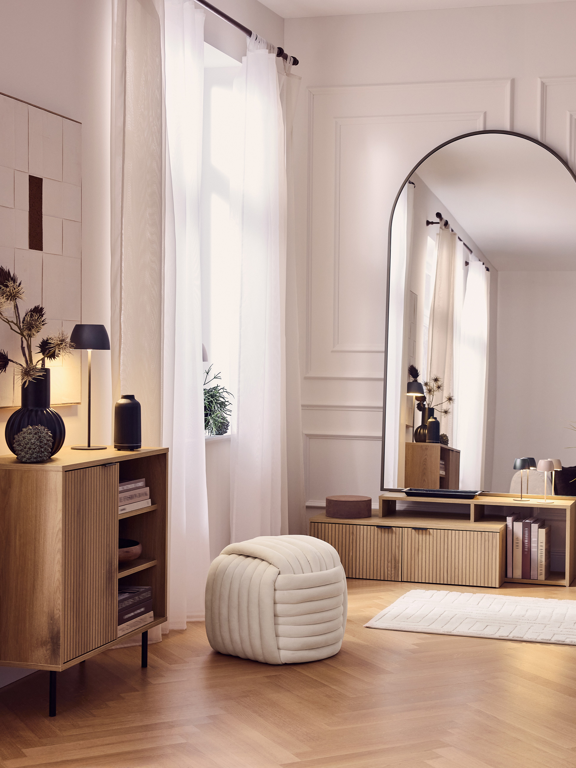 Salon moderne avec des armoires en bois, un grand miroir, un pouf et des rideaux blancs.