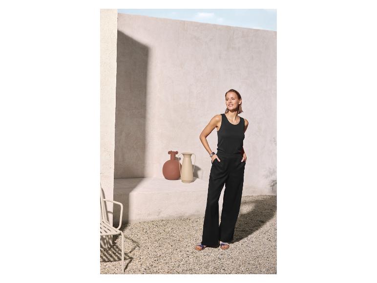 Femme en débardeur noir et pantalon large noir, souriante dans une cour ensoleillée.