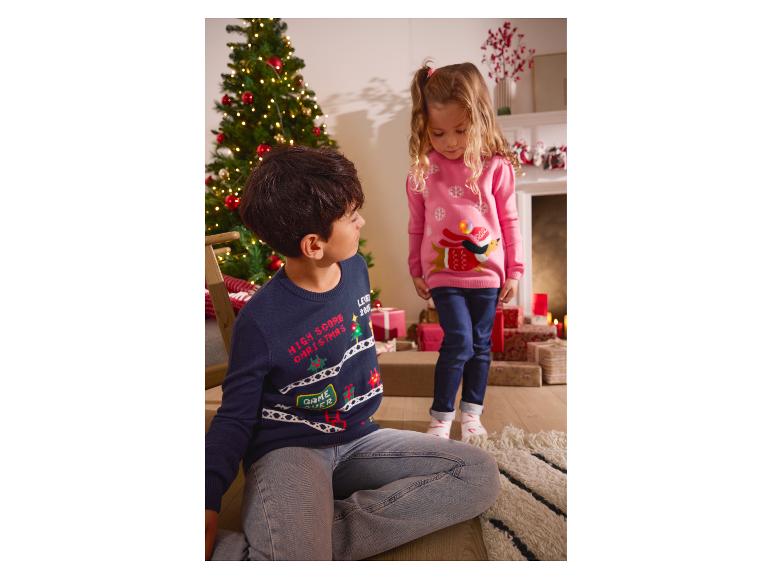 Deux enfants en pulls de Noël, un garçon au sol et une fille debout, à côté d'un sapin de Noël.