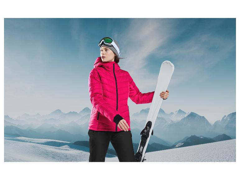 Une femme en veste de ski rose et masque de ski tient des skis blancs dans les montagnes.