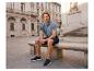 Homme en polo bleu et short assis sur un banc en pierre sur une place de ville.