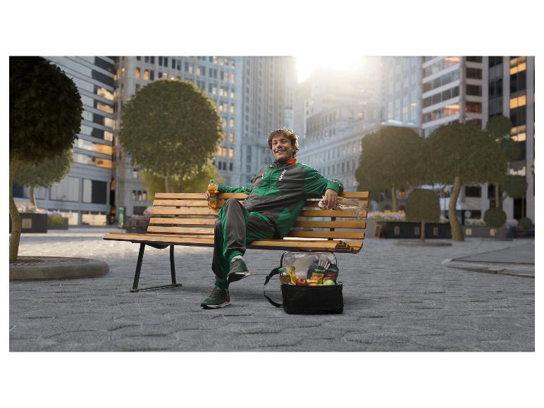 Homme en survêtement sur un banc de parc avec un sac isotherme rempli de fruits frais et une boisson.