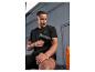 Homme en tenue de sport noire utilisant un pistolet de massage sur son bras, avec une bouteille d'eau orange à proximité.