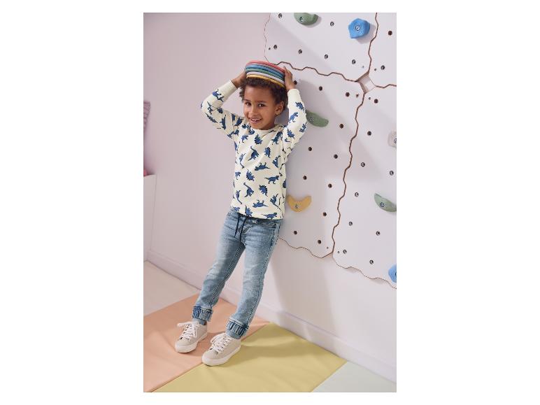 Un garçon souriant en haut à motifs de dinosaures et jean, debout près d'un mur d'escalade.