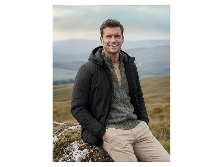 Homme en veste noire et pull vert assis sur un rocher dans un paysage montagneux.