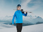 Un garçon en tenue de ski bleue portant des skis sur son épaule, devant un paysage de montagne enneigé.