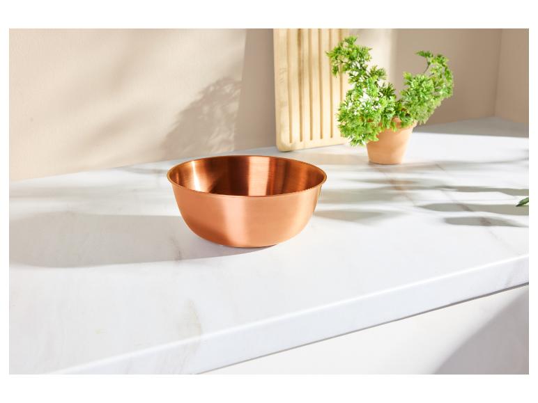 Bol en cuivre sur un comptoir en marbre blanc avec des plantes et une planche à découper en arrière-plan.