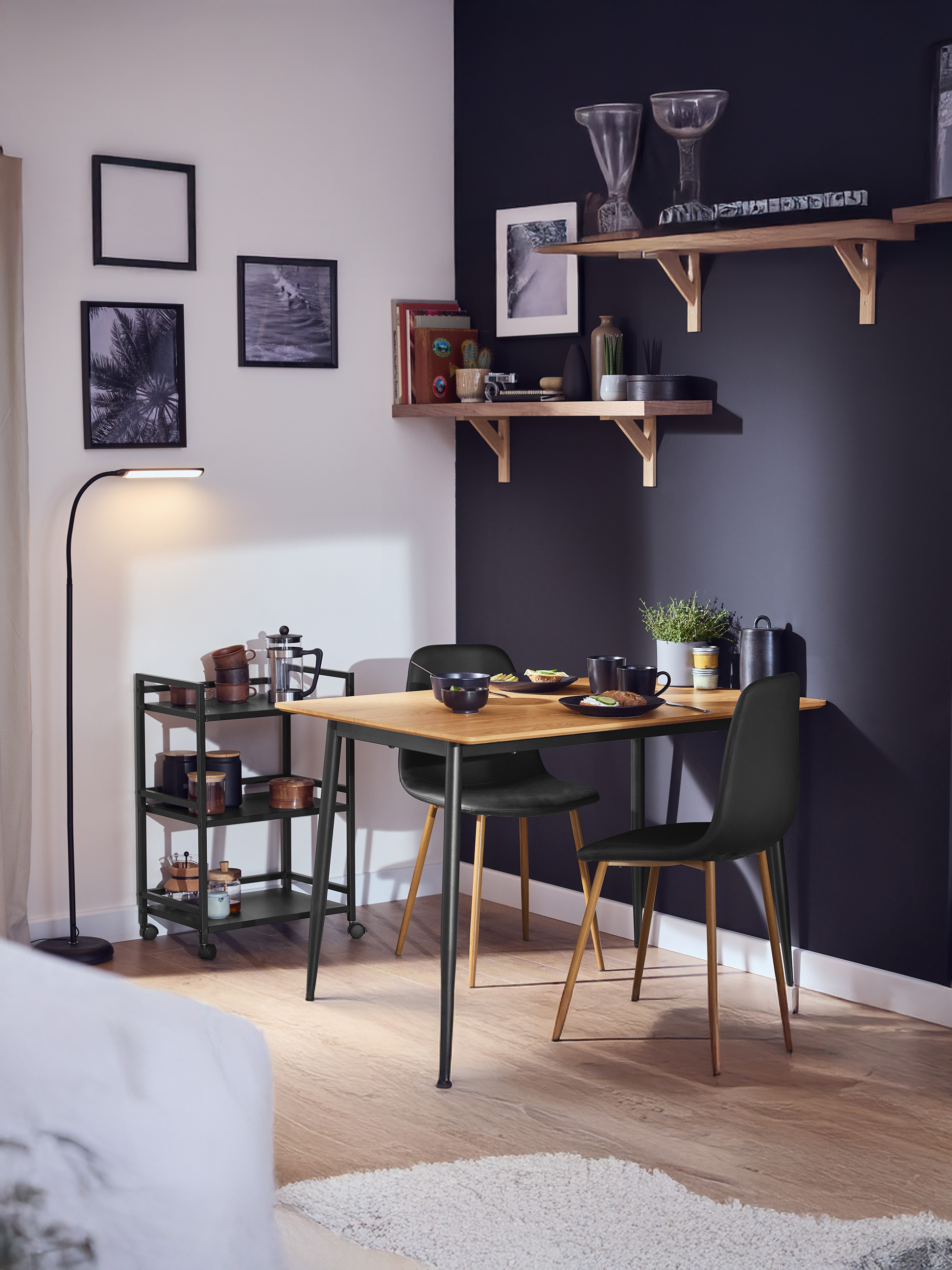Salle à manger avec table en bois, chaises noires, chariot de service et étagères décorées.