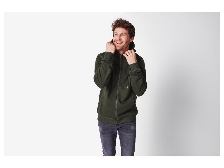 Homme souriant portant un sweat à capuche zippé vert foncé et un jean gris.